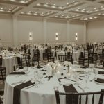 Elegant ballroom reception setup with round tables, dark chiavari chairs, white linens, and modern column lighting