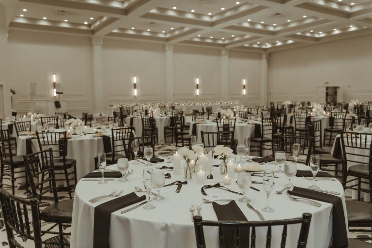 Elegant ballroom reception setup with round tables, dark chiavari chairs, white linens, and modern column lighting