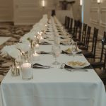 Long wedding reception table with white linens, dark chiavari chairs, white rose centerpieces, and candlelit place settings