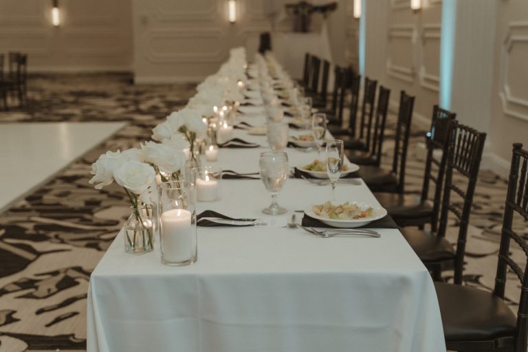 Long wedding reception table with white linens, dark chiavari chairs, white rose centerpieces, and candlelit place settings