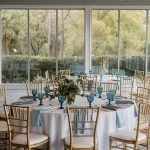 Elegant reception tables with gold chiavari chairs, blue glassware, and garden view through floor-to-ceiling windows