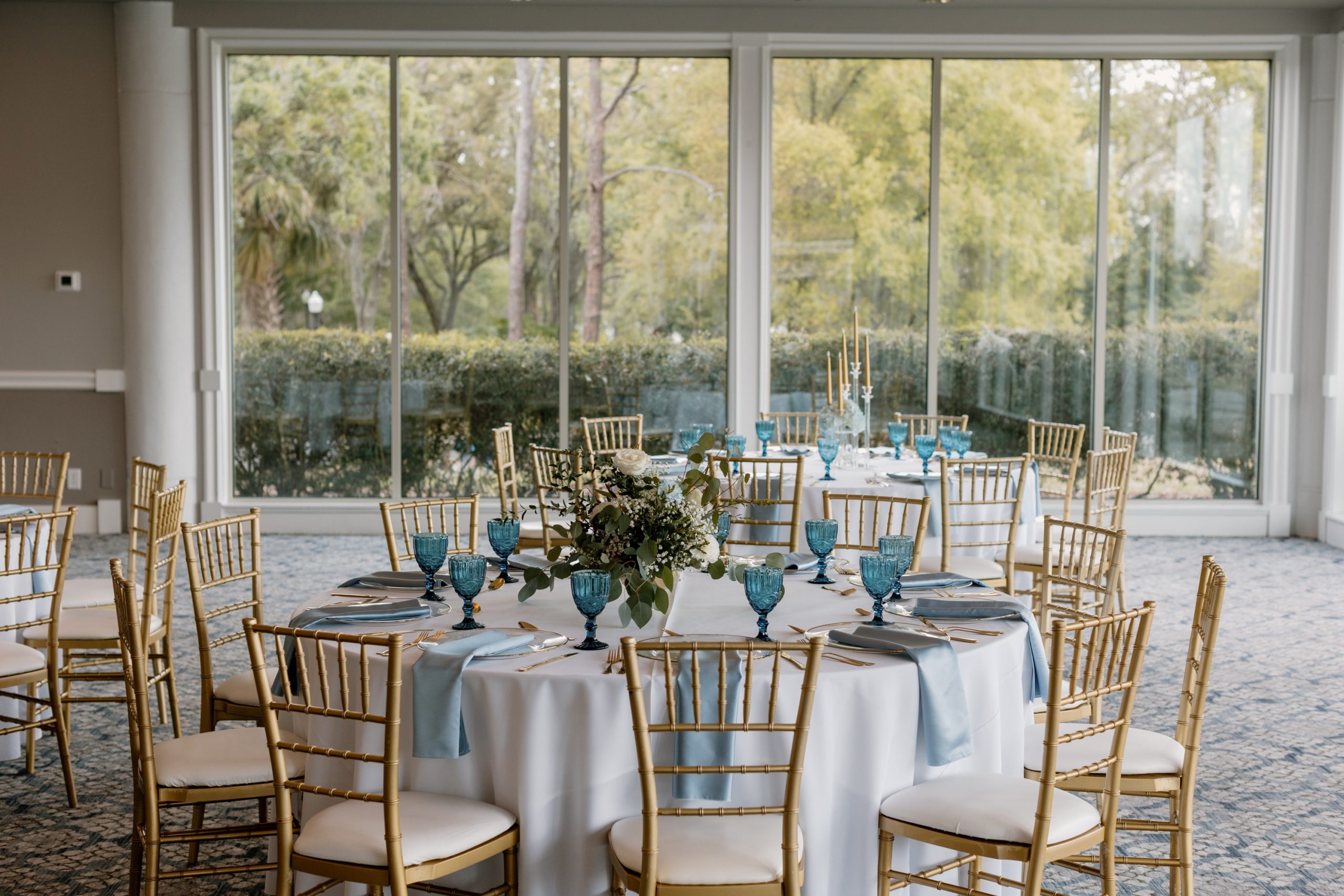 Elegant reception tables with gold chiavari chairs, blue glassware, and garden view through floor-to-ceiling windows