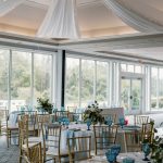 Elegant wedding reception setup with gold chiavari chairs, blue linens, and tall floral centerpieces under a draped tent ceiling