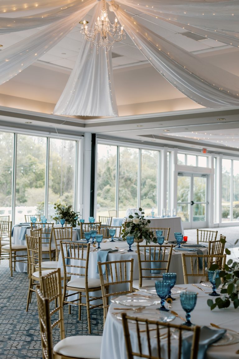Elegant wedding reception setup with gold chiavari chairs, blue linens, and tall floral centerpieces under a draped tent ceiling