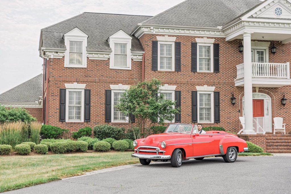 Newlyweds ride in classic red convertible past brick estate at King Cole Farm wedding venue