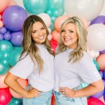 Two women in white tees smile in front of colorful balloon arch in coral, purple, teal, and cream