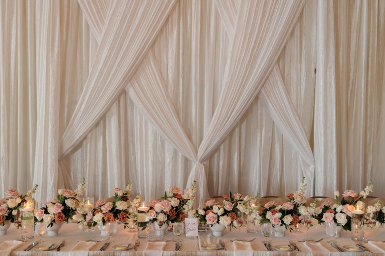 Elegant head table with ivory draping backdrop, blush and coral rose centerpieces, and candlelight