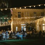 Evening outdoor wedding reception with string lights illuminating rustic building and guests seated at tables