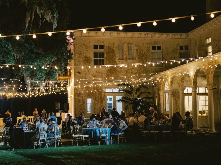 Evening outdoor wedding reception with string lights illuminating rustic building and guests seated at tables