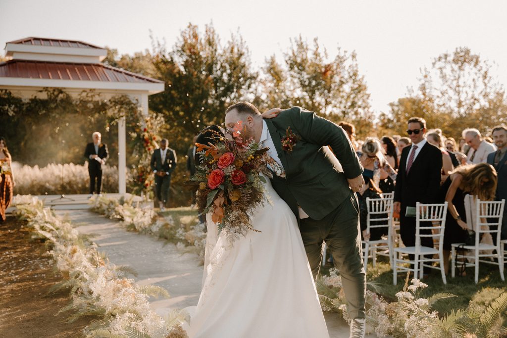 Newlyweds share first kiss walking down ceremony aisle lined with pampas grass and autumn blooms