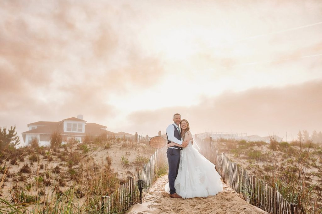 Bride and groom embrace on beach boardwalk with coastal home visible behind sand dunes at sunset