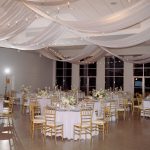 Elegant wedding reception with gold chiavari chairs, white linens, floral centerpieces, and draped ceiling fabric with string lights