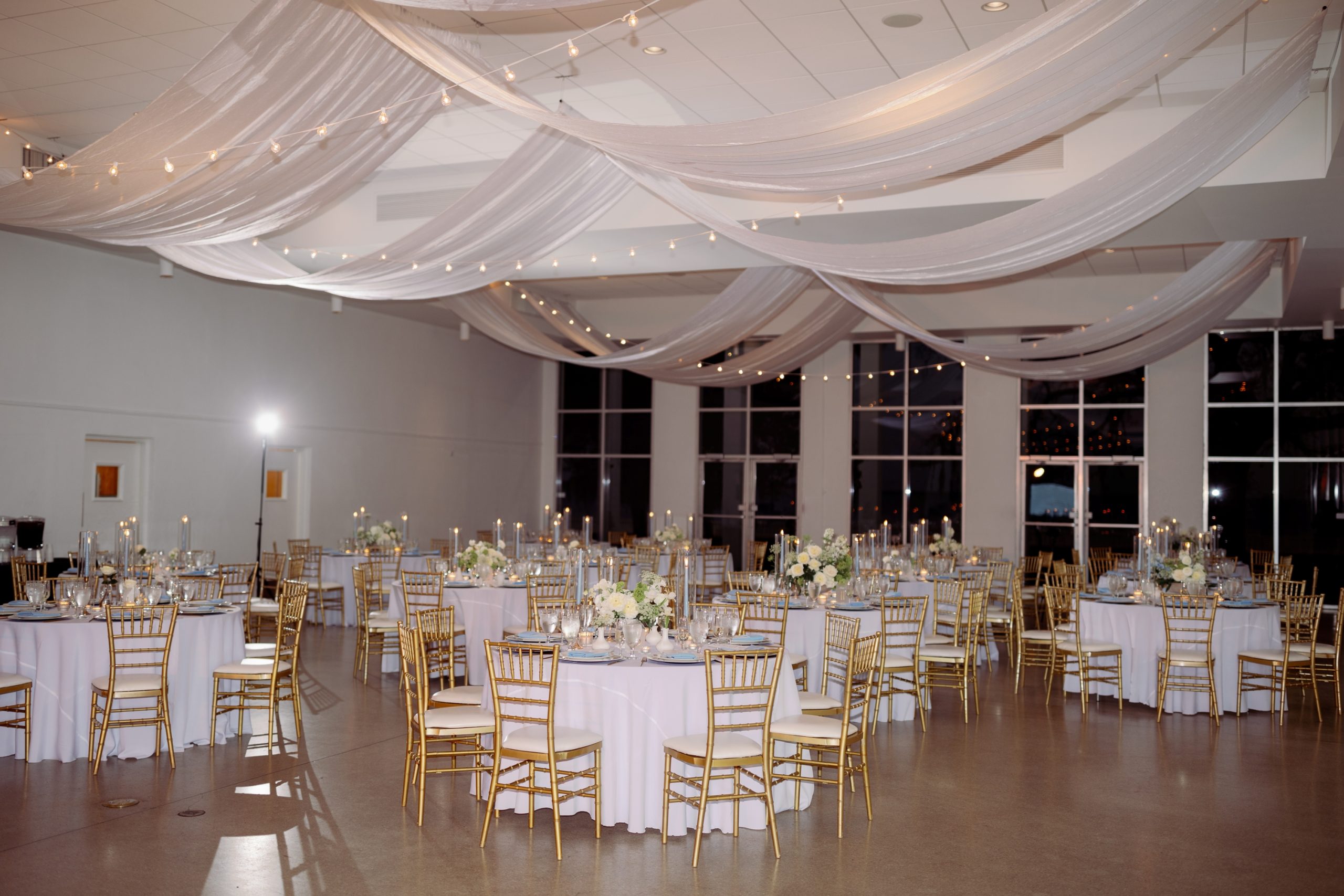 Elegant wedding reception with gold chiavari chairs, white linens, floral centerpieces, and draped ceiling fabric with string lights