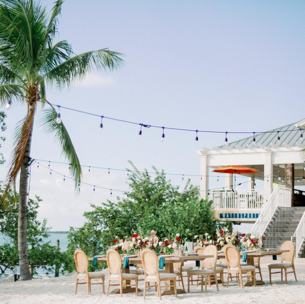 Waterfront reception dining setup with floral centerpieces under string lights and palm trees