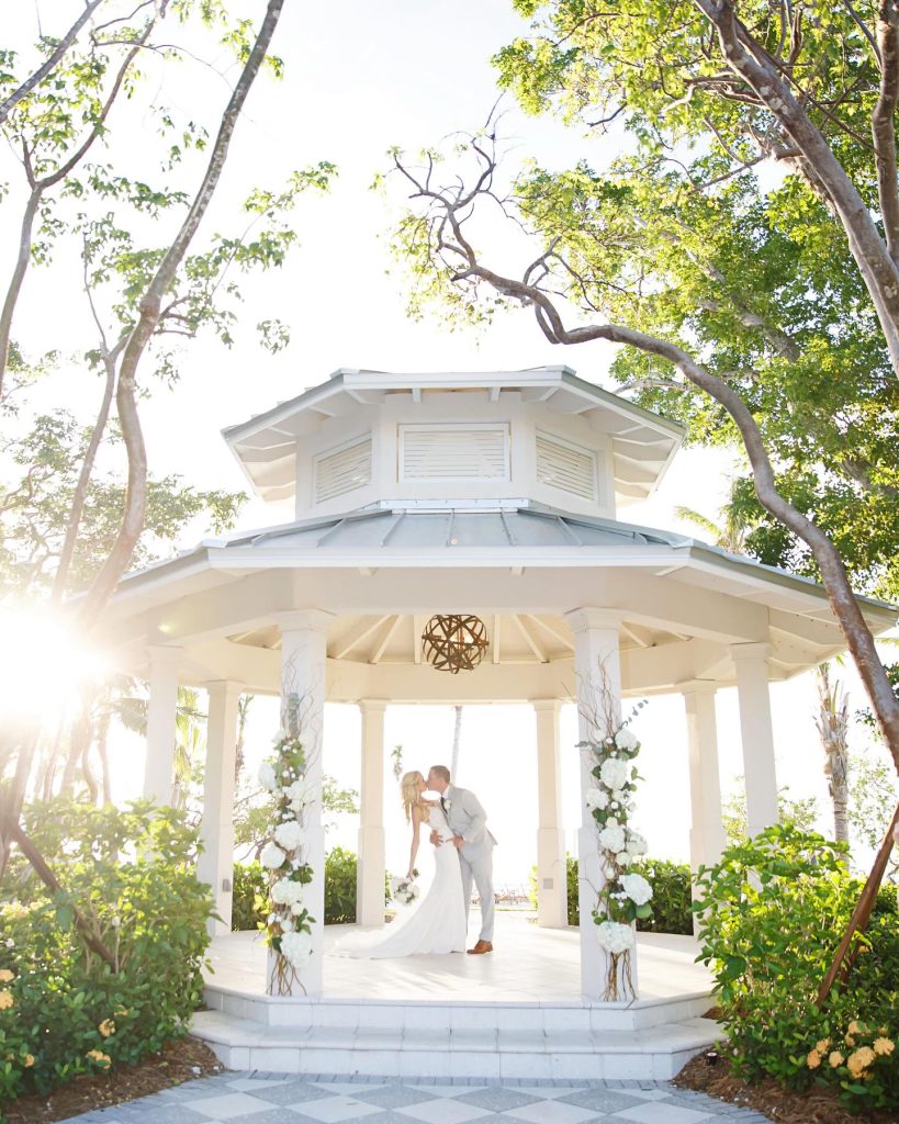 Newlyweds embrace under white gazebo adorned with white blooms and greenery surrounded by tropical foliage