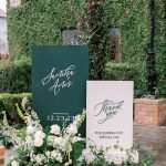 Custom calligraphy wedding signs with white flowers and greenery against ivy-covered wall backdrop