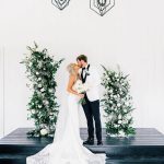 Bride and groom kiss between lush green and white floral pillars under geometric chandeliers