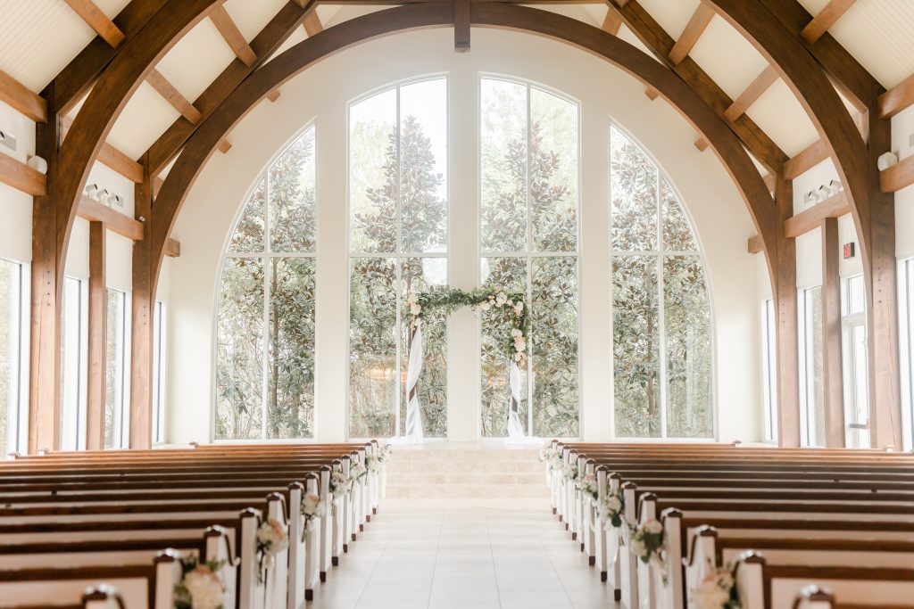 Chapel interior with exposed wood beams, arched windows, and floral ceremony arch framing the aisle