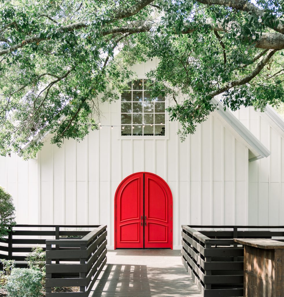 White barn venue with distinctive red arched doors framed by overhanging oak trees at The Carriage House