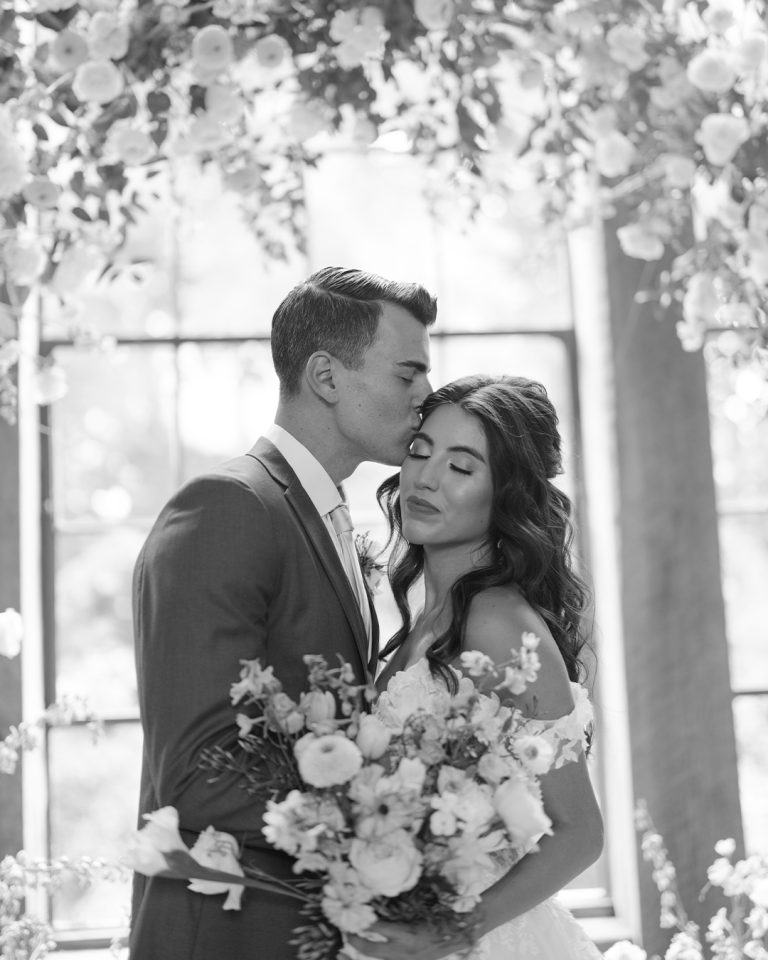 Groom kisses bride's forehead as she holds lush bouquet in black and white portrait under blooming trees