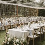 Outdoor reception with cross-back chairs, white linens, lush floral arrangements, and cascading string lights overhead