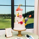 Three-tier white wedding cake decorated with fresh pink, yellow, and purple blooms on gold stand