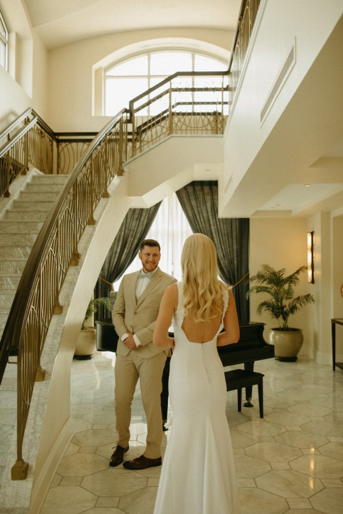 Groom in tan suit greets bride in backless gown in elegant foyer with curved staircase