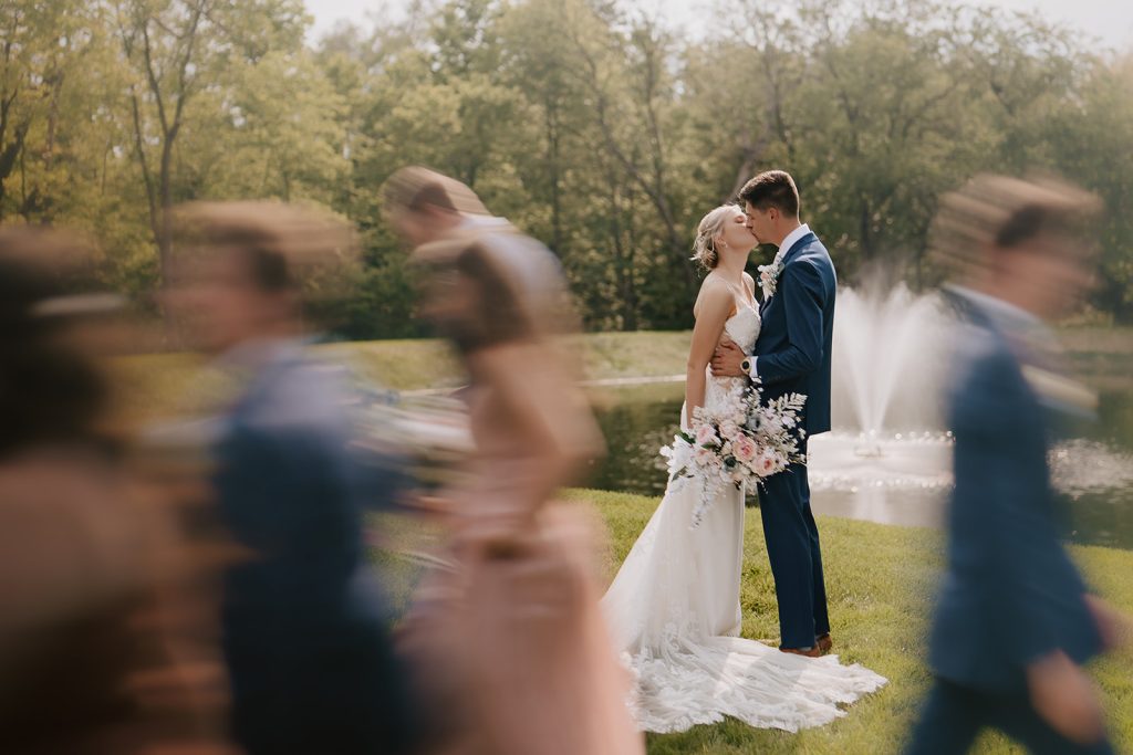 Couple shares first kiss as newlyweds by pond with guests in foreground