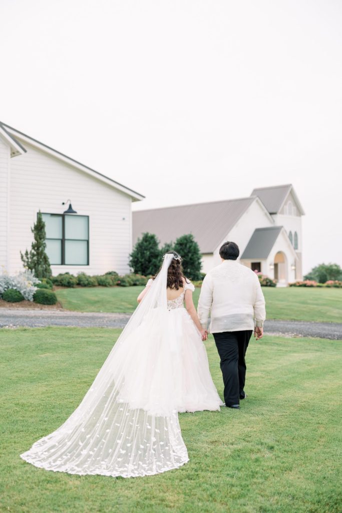 Bride in flowing veil and ballgown walks hand-in-hand with groom across green lawn toward white chapel buildings