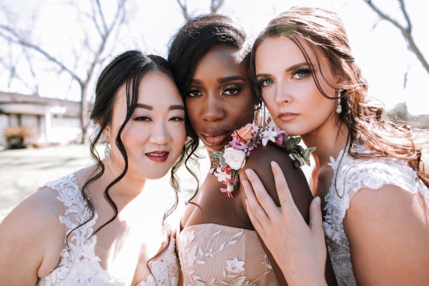 Three brides in lace gowns pose together wearing soft updo hairstyles and fresh floral accessories