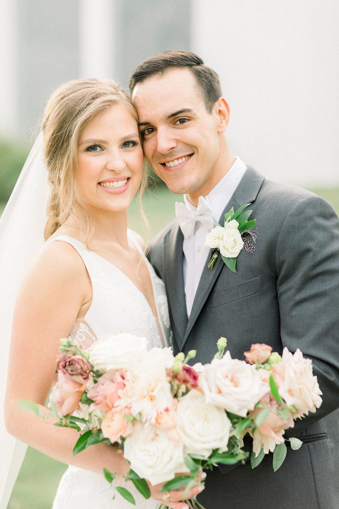 Smiling newlyweds pose together, bride in veil with pink and white bouquet, groom in gray suit