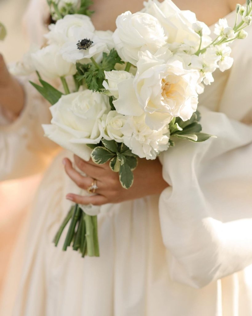 Bride holding elegant white and cream bouquet with greenery in flowing wedding gown