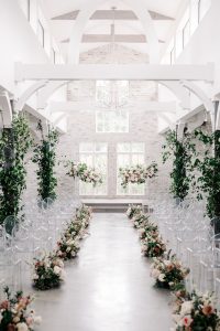 Modern white chapel ceremony space with exposed beams, clear ghost chairs, and pink floral aisle arrangements