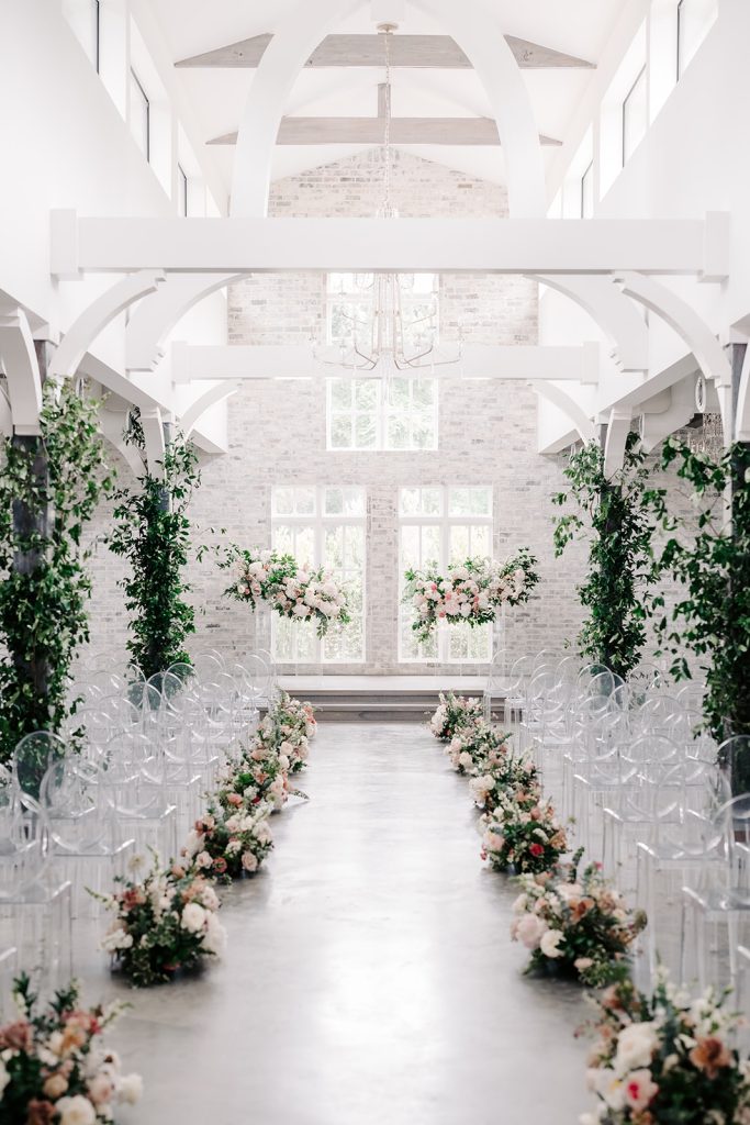 Modern white chapel ceremony space with exposed beams, clear ghost chairs, and pink floral aisle arrangements