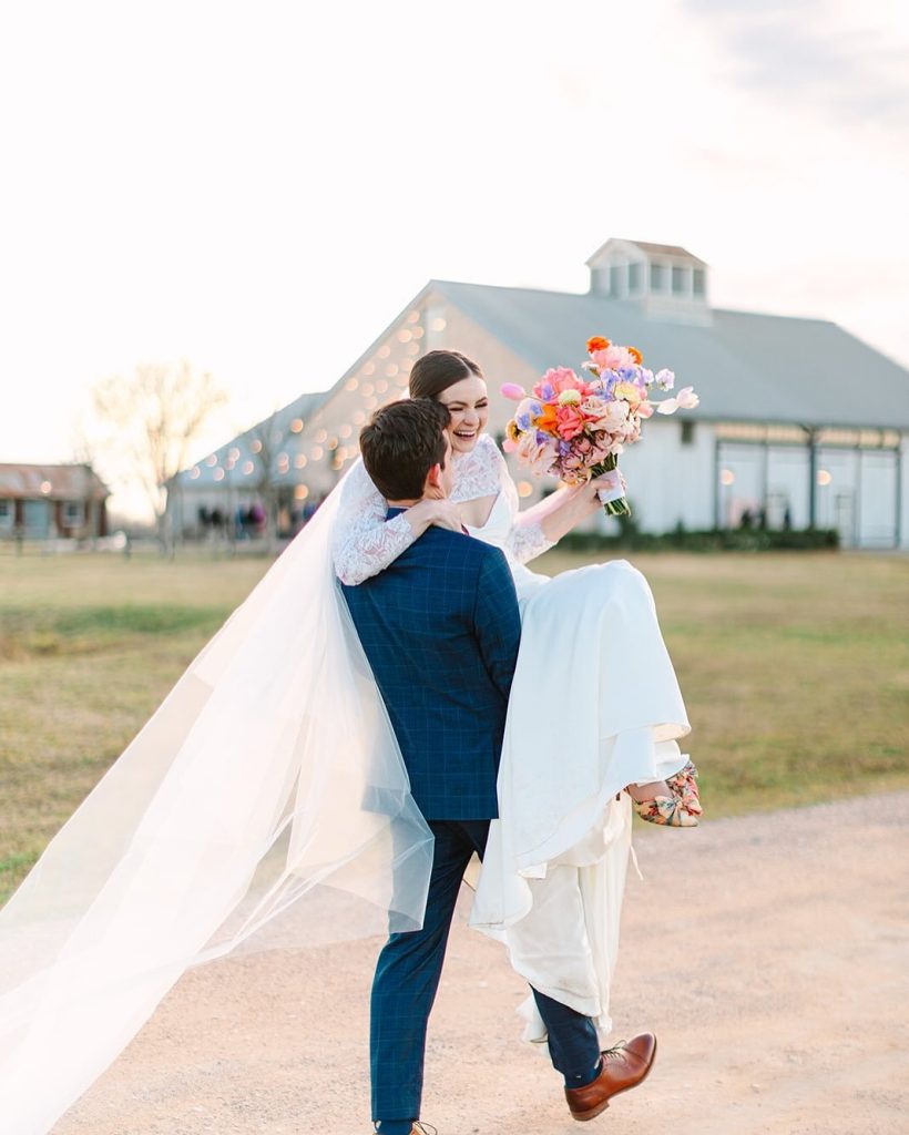 Groom lifts bride holding colorful bouquet outside modern barn venue at sunset