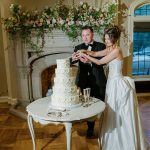 Bride and groom cutting tiered white wedding cake in front of fireplace adorned with lush floral arrangements