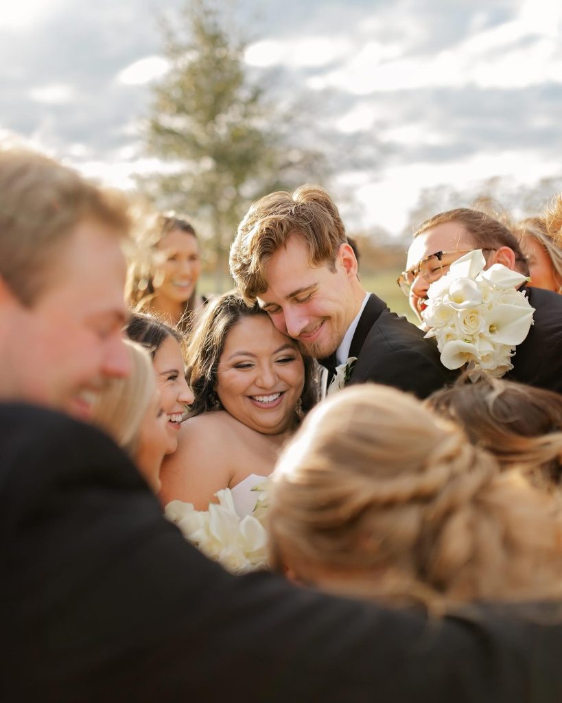 Newlyweds embrace surrounded by joyful wedding party holding white bouquets outdoors