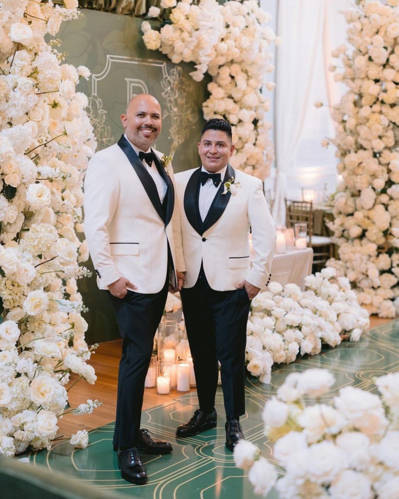 Two grooms in white tuxedo jackets standing beneath luxurious white floral ceremony arch