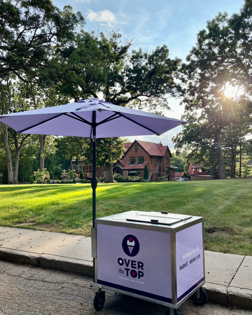 Over the Top ice cream cart with purple umbrella positioned on lawn in front of historic brick building