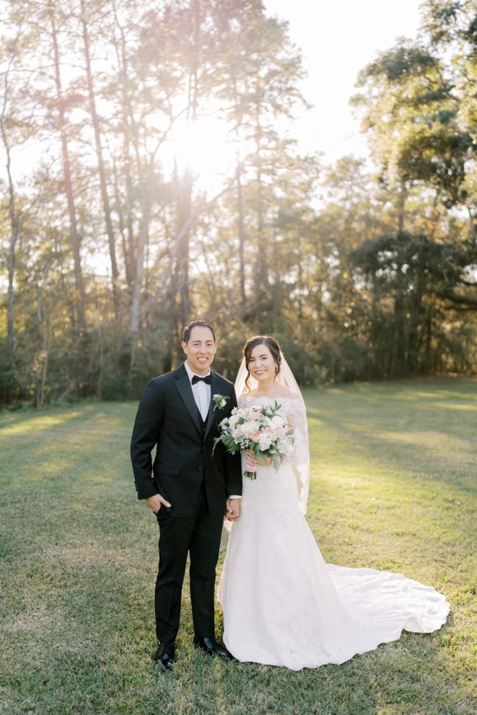 Bride and groom stand in sunlit garden clearing surrounded by trees