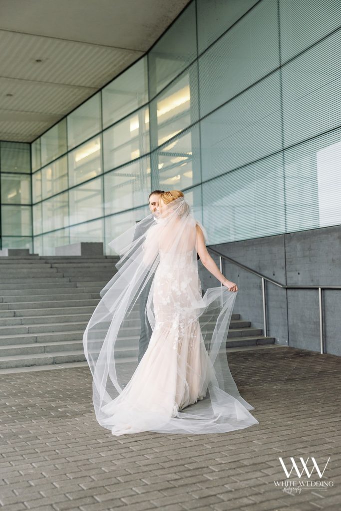 Bride in lace gown with cathedral veil flowing dramatically at modern glass building