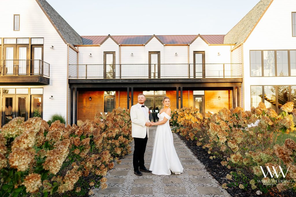Bride and groom pose in front of modern farmhouse venue with autumn landscaping and second-story balcony
