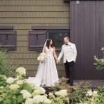 Newlyweds holding hands outside rustic barn venue surrounded by white hydrangea blooms in Indianapolis