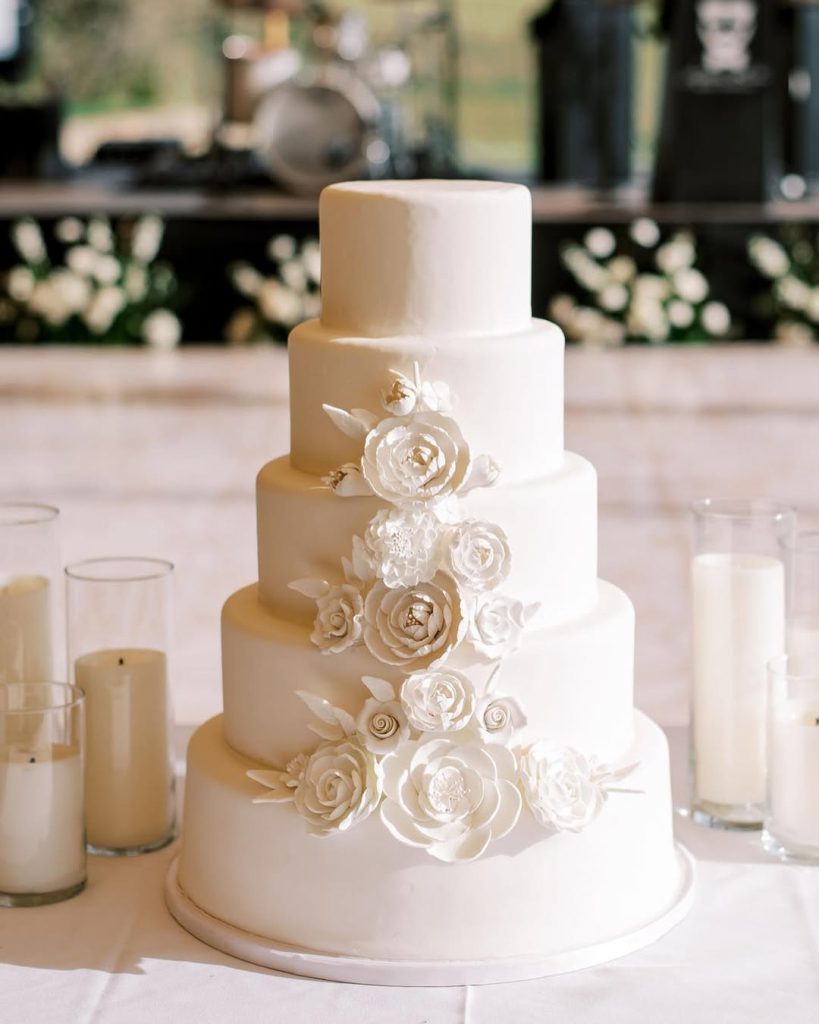 Five-tier white wedding cake adorned with cascading sugar flowers and gold accents, surrounded by pillar candles