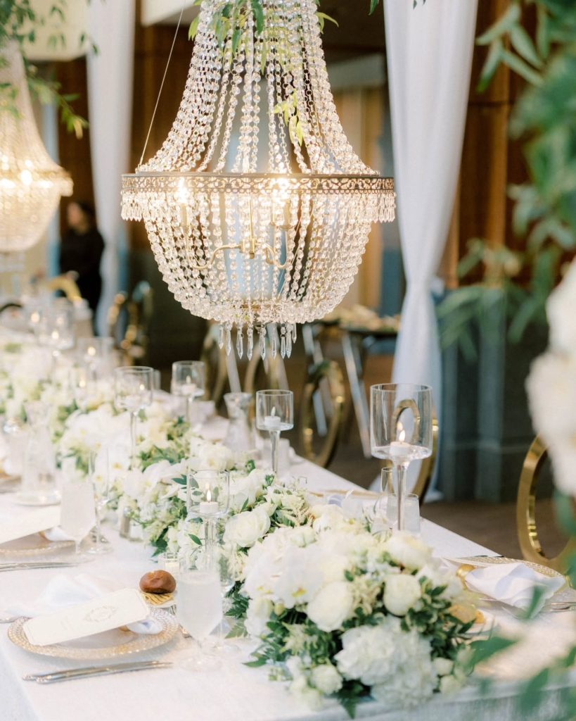Crystal chandelier centerpiece suspended above white-linened wedding table with gold chargers, white floral garland, and glass candle holders