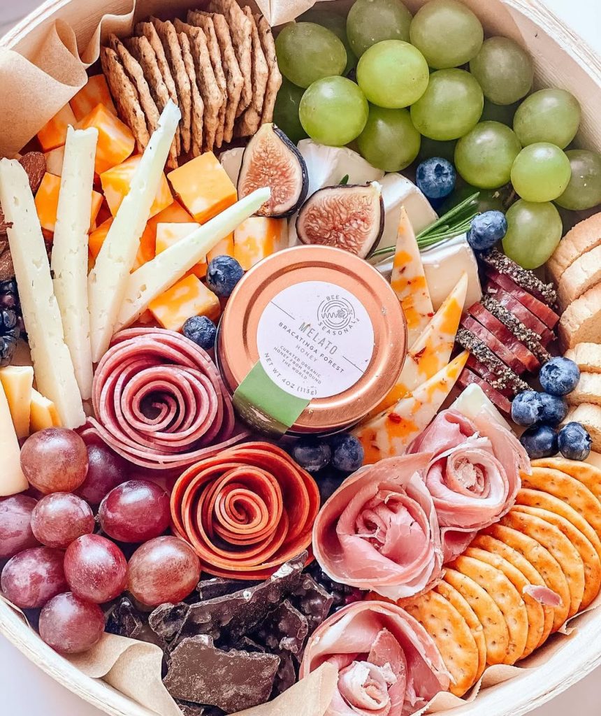 Round charcuterie box filled with crackers, cheeses, grapes, figs, and decorative salami roses