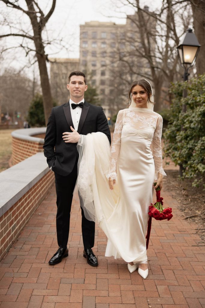 Newlyweds walk arm-in-arm through urban park with bride holding red bouquet