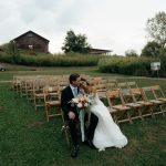 Bride and groom embrace at outdoor ceremony with wooden folding chairs and rustic barn backdrop