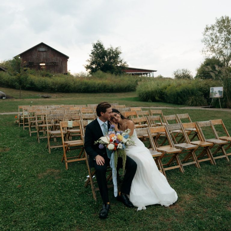 Bride and groom embrace at outdoor ceremony with wooden folding chairs and rustic barn backdrop