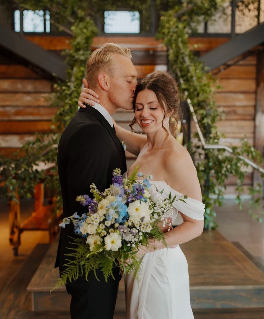Newlyweds share an intimate moment in a rustic barn venue surrounded by lush greenery and a colorful wildflower bouquet